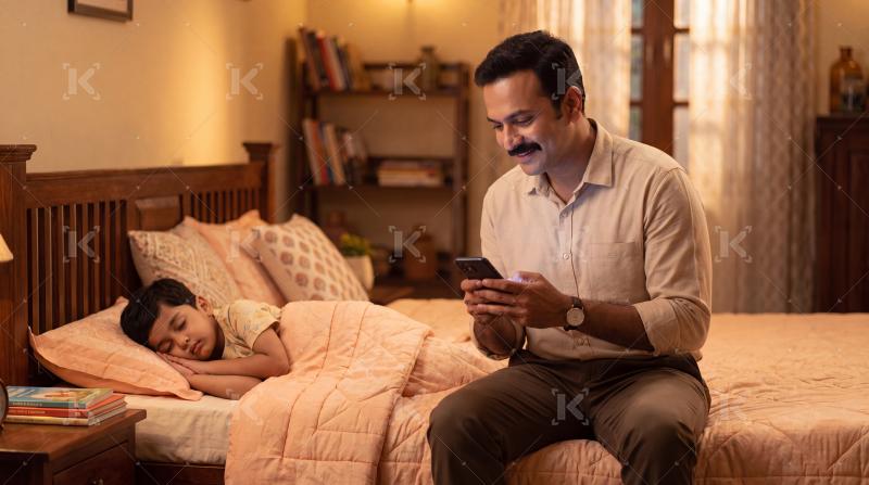 Happy indian father sitting on bed using smartphone