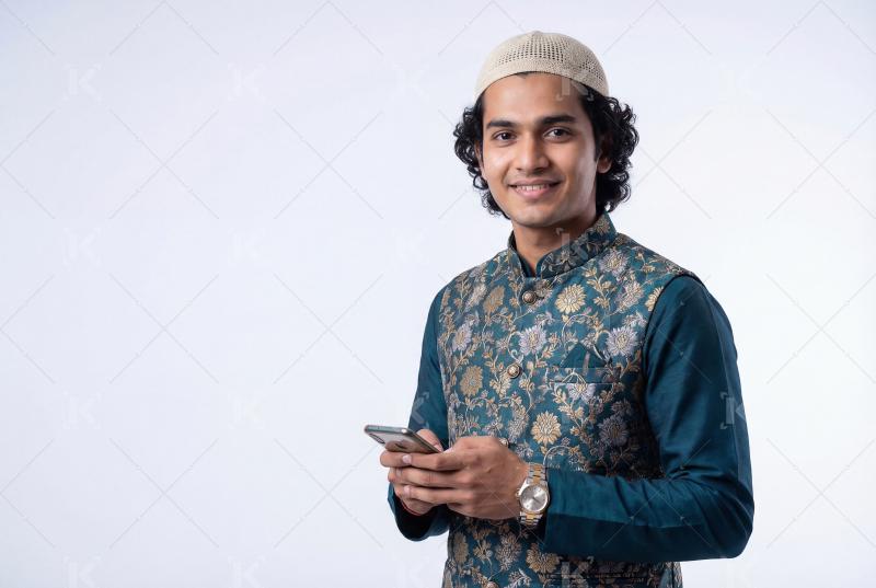 A smiling young Muslim man in traditional attire using a smartph