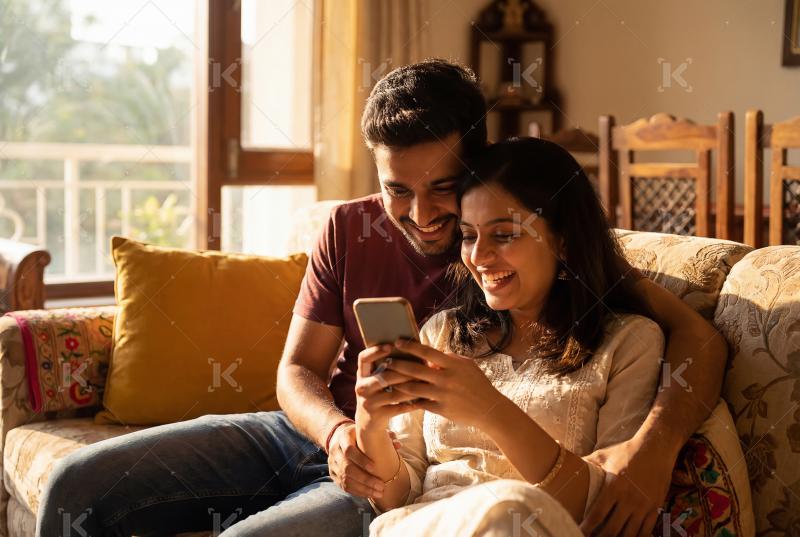 Young indian couple sitting on sofa using smartphone together