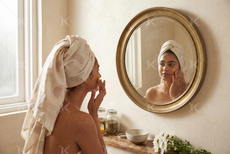 A woman wrapped in a towel gently applies skincare while looking