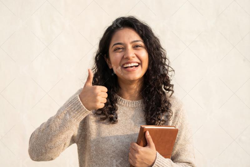 Young indian female college student holding book showing thumbs up standing on white background