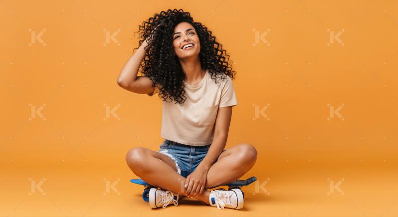 Joyful Woman with Curly Hair Sitting on Skateboard, Orange Backg