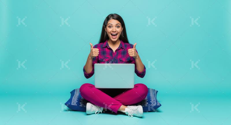 A young woman sits cross-legged on a cushion with a laptop, givi