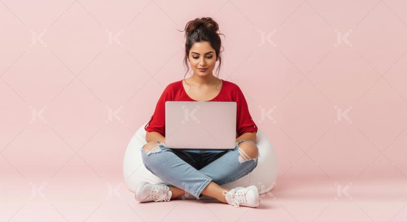 A young woman sits cross-legged on a gray bean bag with laptop