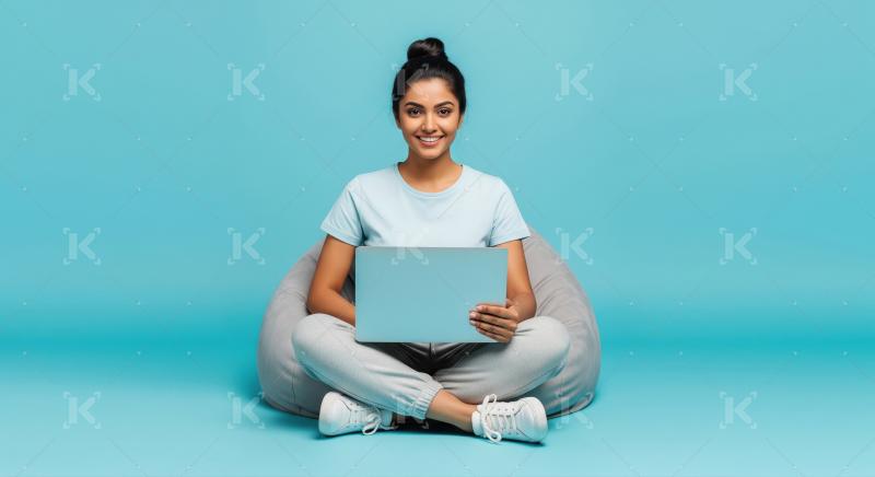 A young woman sits cross-legged on a gray bean bag with laptop