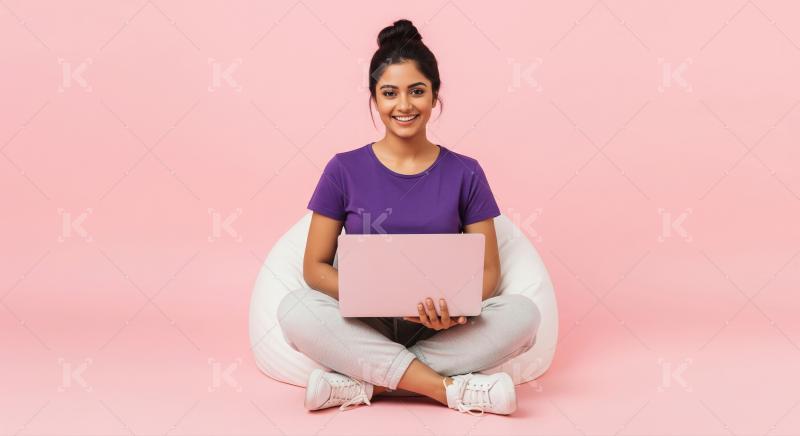 A young woman sits cross-legged on a gray bean bag with laptop
