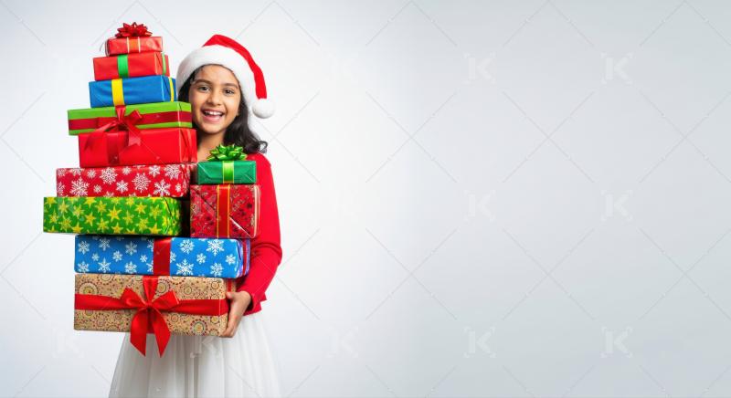 A smiling Indian girl in a Santa hat holds a stack of Christmas presents wrapped with red ribbons