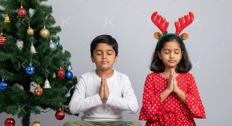 A little boy and girl sit peacefully in prayer pose beside a decorated Christmas tree