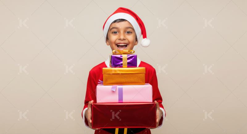 A joyful Indian boy in a Santa hat holds a colorful stack of Christmas presents, beaming with excitement