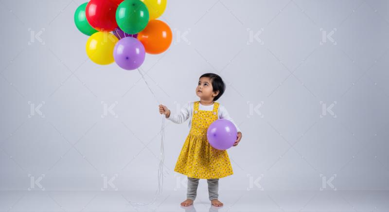 A young child in a yellow dress stands barefoot indoors, holding colorful balloons and a purple ball against a plain background.