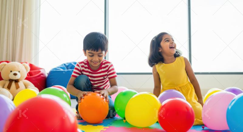 Two children, surrounded by colorful balloons, play together on the floor in a bright indoor setting