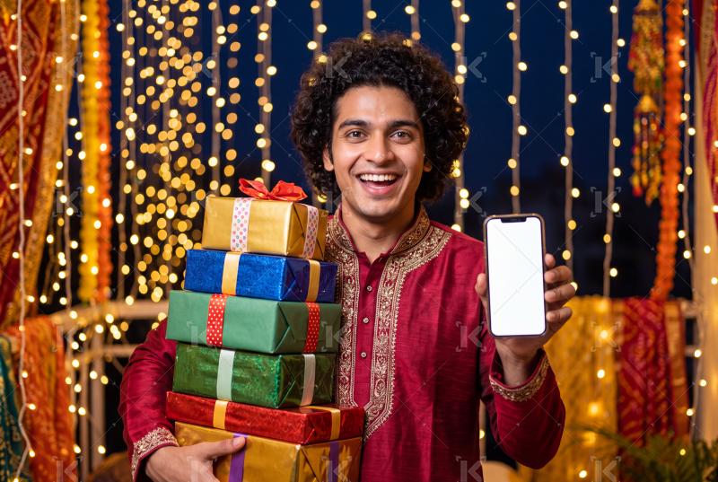 Young indian man holding gift boxes and showing smartphone screen on traditional diwali festival